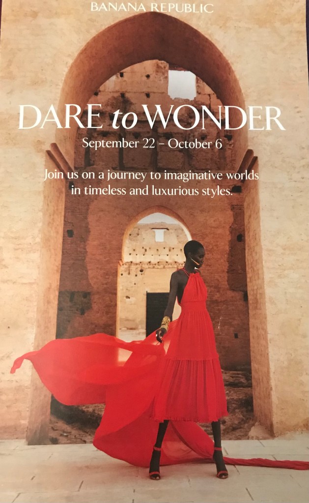 The cover of a Banana Republic advertisement. A woman of African descent wears a dramatic red dress with a halter top and a flowing scarf behind her. She wears red high-heeled sandals. She stands in front of beige sandstone walls and an arched doorway. The white text reads: Dare to Wonder. Join us on a journey to imaginative worlds in timeless and luxurious styles.