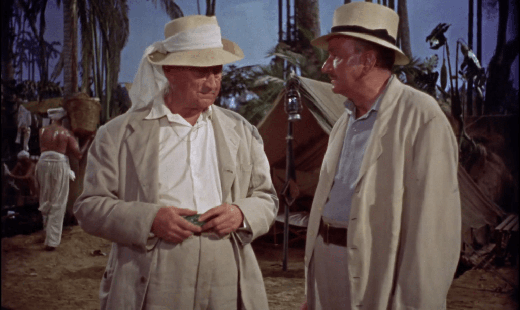 Screenshot from a scene in the desert. Two late middle-age white men stand in the foreground. They were khaki linen suits over white shirts and beige or khaki hats. They are sweaty and slightly dirty. Behind them are khaki tents. Shirtless, sweaty men who are meant to be Egyptian laborers wear white headcoverings and khaki linen pants. They carry baskets and tools. There are a few palm trees in the background of the image.