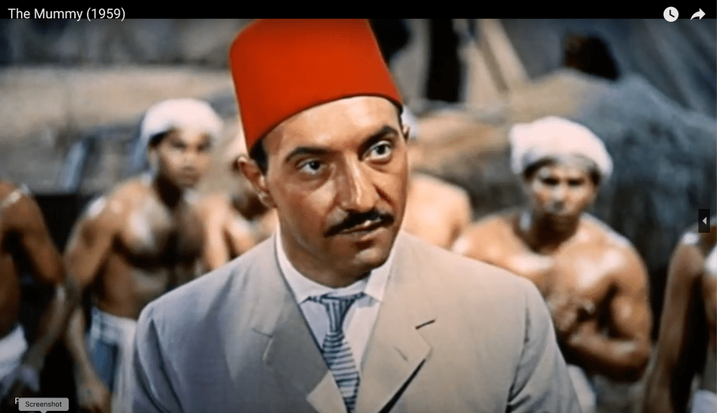 A man of Greek descent wearing heavy brown makeup to emphasize his foreign-ness, stands in the center of the screenshot. He wears a crisp beige suit over a white shirt and blue and white striped necktie. On his head, he wears a red Fez. In the background, sweaty shirtless men wear white head coverings and khaki linen pants. They are in the desert at the site of an archaeological dig.