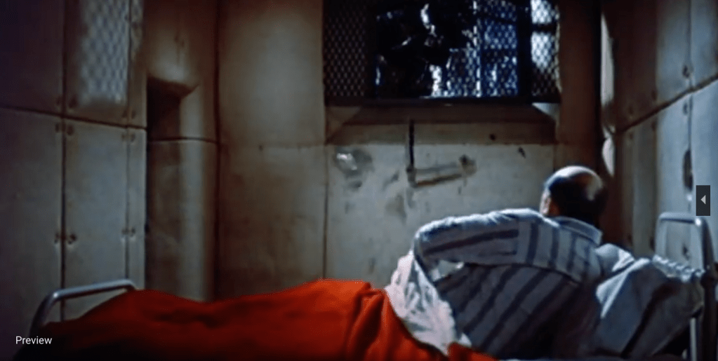 The lower third of the image shows an old white man in bed. He wears grey and white pajamas. The walls of the room are offwhite and padded. The window is barred and occupies the upper two-thirds of the image. An ominous figure is breaking through the bars. On the bed is a bright red blanket, the only bright color in the otherwise drab and shadowy room.